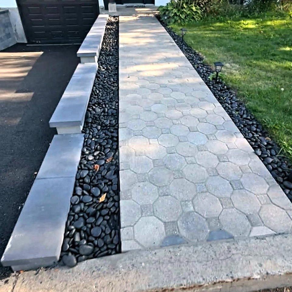 Allée résidentielle en pavé uni gris, bordée d’un lit de galets noirs et de larges dalles de béton, menant à la porte d’entrée d’une maison blanche.